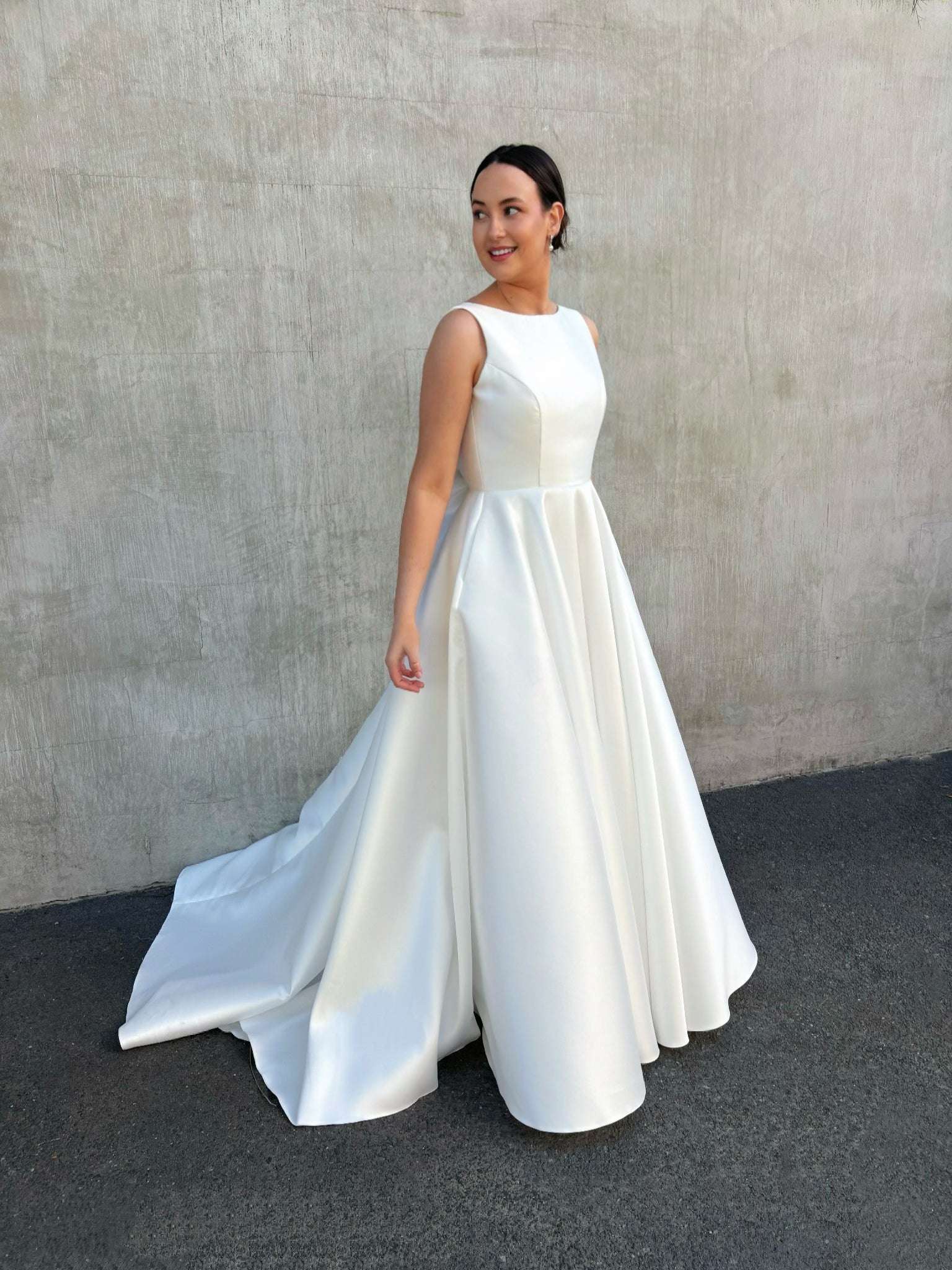 Side profile of boat-neck wedding gown showing the structured waist, flowing full skirt and train, and glossy Mikado finish.