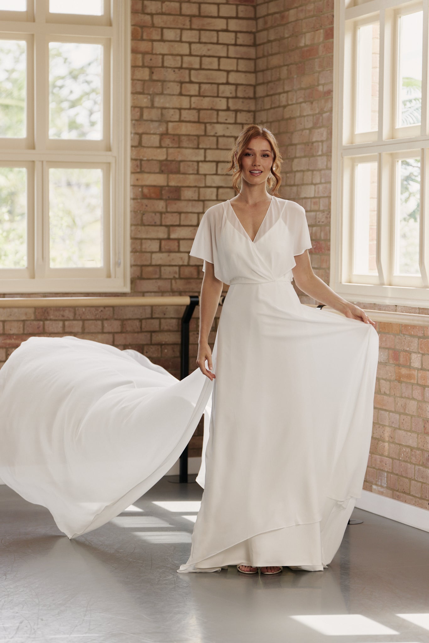 Bride wearing Clare wedding dress with flutter sleeves, V-neck crossover bodice, and flowing A-line skirt with train.